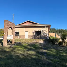 This image shows the exterior of 3 Brothers Painting & Home Improvements, LLC, featuring a tan building with brick accents, a custom brick arch sign, and a sunny blue sky.
