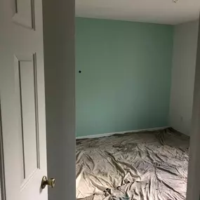 An interior room is mid-renovation by 3 Brothers Painting & Home Improvements, LLC. A freshly painted teal wall contrasts with white trim, and protective drop cloths cover the carpeted floor.