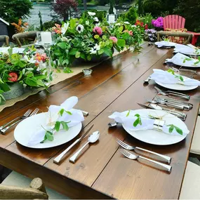 A beautifully styled outdoor dining table set for a private event catered by Chef Joe, featuring elegant white plates, polished silverware, and neatly folded napkins adorned with fresh greenery. The rustic wooden table is decorated with vibrant floral centerpieces and surrounded by lush garden scenery, creating a warm and inviting atmosphere. This setup reflects Chef Joe’s ability to transform any space into a restaurant-quality dining experience for intimate gatherings and special events throug
