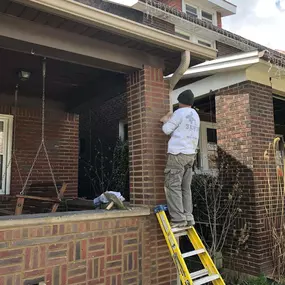 A construction worker on a yellow ladder repairing brickwork on a porch column of a red brick house with decorative patterns.