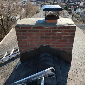 A newly repaired brick chimney with a black metal cap on a shingled rooftop, with ladders and a cityscape in the background.