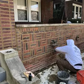 Two workers repairing a decorative brick porch wall; one squatting with mortar, the other on a ladder restoring the brick column.