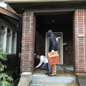 Two workers restoring a brick porch entrance; one kneeling to repair the wall, the other holding an orange crate, standing nearby.
