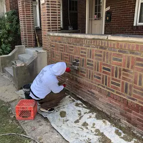 A worker in a white hoodie and red gloves repairs a decorative brick wall, applying mortar while kneeling on a tarp-covered ground.