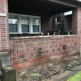 A completed brick wall repair on a front porch, showing restored decorative brick patterns. Construction tools and debris remain nearby.