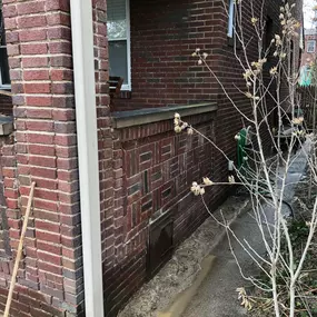 A repaired brick wall on a porch corner with intricate brick patterns. A downspout, broom, dried plant, and wet sidewalk visible.