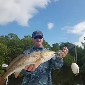 A man in a blue camo shirt and sunglasses holds a large fish, possibly a redfish, on a boat. Lush green trees and a blue sky with white clouds are in the background.