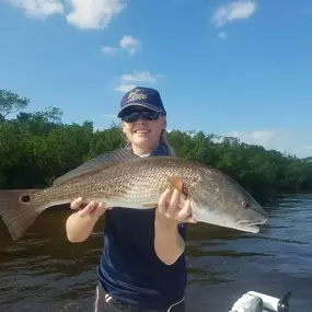 A woman in a navy shirt, shorts, sunglasses, and a cap holds a large redfish on a boat. The fish is displayed horizontally, and the background shows water, trees, and a blue sky with clouds.