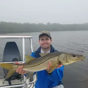 A smiling man on a boat holds a large snook fish horizontally. He wears a blue jacket, shorts, and a camouflage cap. The background shows a foggy, overcast day over water.