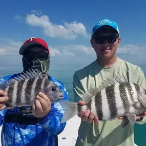 Two men on a boat hold up their catches, striped sheepshead fish. One man wears a red cap and face covering, the other a blue cap and sunglasses. A blue sky and ocean are visible.