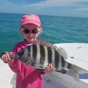 A young girl in pink clothing and sunglasses holds a striped sheepshead fish on a boat. The ocean and a blue sky are visible in the background. She appears to be smiling and proud of her catch.