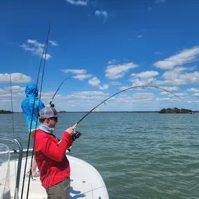 Two people fish from a white boat on a sunny day with a blue sky and scattered clouds. One person in red actively reels in a line, while another in blue stands in the background. Water and distant land are visible.