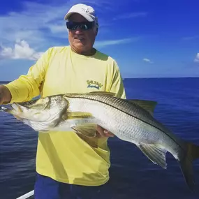 A man in a yellow shirt and sunglasses holds a large snook fish on a boat. The fish is silver and elongated.  The background shows a blue sky with clouds and a dark blue ocean.