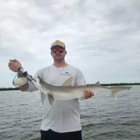 A man in a light grey shirt, sunglasses, and a cap holds a small shark on a boat. The shark is light grey and white.  The background shows water and a cloudy sky.