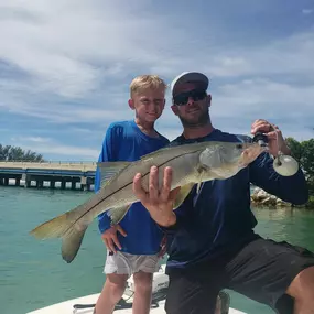 A man and a young boy stand on a boat, holding a large snook fish. The man kneels, showing the fish to the boy.  A bridge and clear blue water are in the background under a partly cloudy sky.