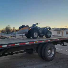 A black ATV with a snowplow attachment is secured on a flatbed tow truck at sunset in a small-town setting.