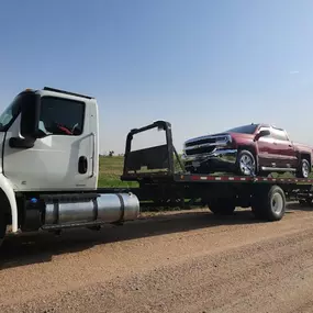 A white tow truck hauling a red pickup with front-end damage on a flatbed trailer along a rural dirt road.