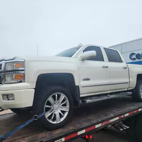 A white Chevrolet Silverado pickup truck secured on a flatbed tow truck outside a dealership with 