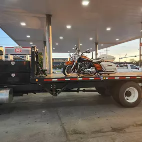 A white flatbed tow truck carrying a classic orange and black touring motorcycle parked at a Phillips 66 gas station during sunset.