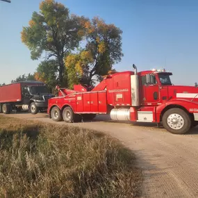 A large red heavy-duty tow truck is towing a black semi-truck with a red container on a rural dirt road.