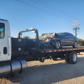A black flatbed tow truck is transporting a heavily damaged dark-colored sedan with a crushed front end on a gravel road.