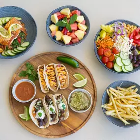 Table with five different dishes containing Tacos, fruit, French fries and salads