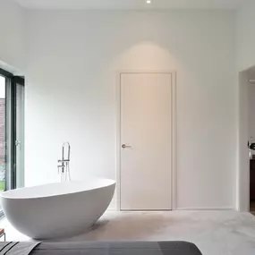 Reduziertes, modernes Interieur mit klarer Formsprache: Ein freistehendes, skulpturales Bad im Schlafzimmer verleiht dem Raum eine besondere architektonische Note. Die bodentiefen Fenster bringen Tageslicht in die minimalistisch gestaltete Umgebung, während warme Lichtakzente eine wohnliche Atmosphäre schaffen. Die Kombination aus Funktion und Design macht den Raum zu einem Statement für zeitgenössisches Wohnen.