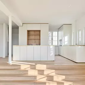 Offene Küche mit zurückhaltender Eleganz: Weiß, Holz und Messing prägen die reduzierte Gestaltung. Die großzügige Raumhöhe und das Tageslicht verstärken die architektonische Klarheit.
