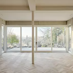 Offener Wohnraum mit raumhoher Verglasung, Sichtbetonwänden und Fischgrätparkett. Architektur, Licht und Landschaft treten in einen ruhigen, atmosphärischen Dialog.