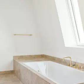 Elegantes Bad mit Einbaubadewanne in Natursteinfassung. Die Kombination aus klarer Formensprache, hellen Materialien und goldenen Armaturen schafft eine luxuriöse, zeitlose Atmosphäre.