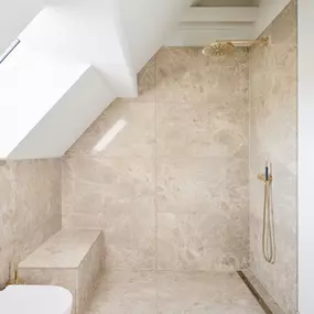 Hochwertig ausgestattetes Duschbad unter der Dachschräge mit Natursteinverkleidung, goldenen Armaturen und integrierter Sitzbank. Eine Kombination aus Funktionalität und eleganter Zurückhaltung.