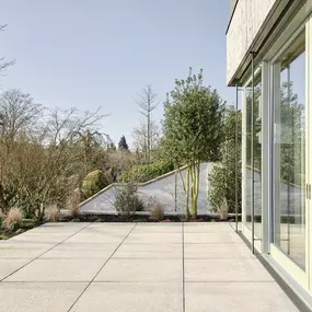 Großzügige Terrasse mit direktem Übergang zur Natur: Die klare Geometrie der Bodenplatten trifft auf weiche Landschaftsformen und üppige Begrünung. Glasfassade und Pflanzung verschmelzen Innen- und Außenraum.