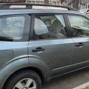 A grey-blue Subaru Forester SUV parked on a residential street. The side and rear windows are clearly tinted, offering increased privacy and a uniform look to the vehicle's glass.