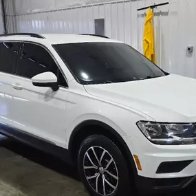 A white Volkswagen Tiguan SUV sits inside a well-lit service bay, likely at Tinted Inc. The vehicle's side and rear windows feature a dark tint, contrasting sharply with the paint.
