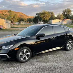 A black Honda Civic sedan parked on a gravel lot outdoors. Its side and rear windows feature a dark tint, adding a sleek aesthetic and enhancing vehicle privacy effectively.