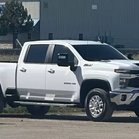 A white Chevrolet Silverado HD pickup truck parked outdoors near a large building. Its side windows feature a dark tint, contrasting sharply with the white paint for enhanced style.