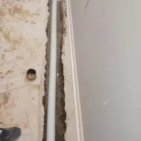 This image shows plumbing work in progress, likely by Sinks & Sewers LLC. A white PVC pipe is laid in a narrow trench dug into a concrete or dirt floor, surrounded by gravel fill. The trench runs alongside a finished interior wall.