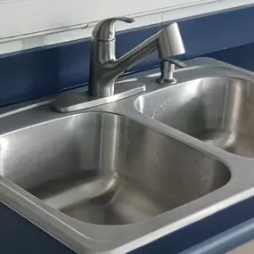 This image showcases a newly installed double-basin stainless steel kitchen sink, likely work done by Sinks & Sewers LLC. A modern brushed nickel faucet is mounted on the sink, set into a dark blue countertop.