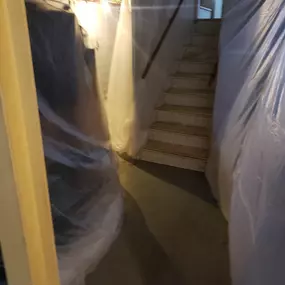 This image shows a basement area prepared for construction or repair work, presumably by Sinks & Sewers LLC. Plastic sheeting covers walls and possibly furniture, protecting them from dust and debris. Stairs lead upwards in the background.