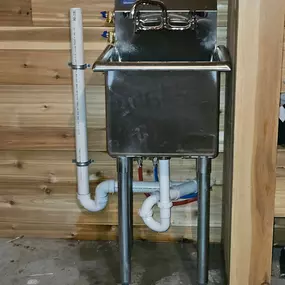 This image shows a freestanding stainless steel utility sink installed against a wood-paneled wall, likely a project by Sinks & Sewers LLC. All plumbing, including supply lines, P-trap, and vent pipe, is exposed.