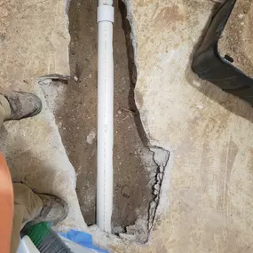 This image depicts plumbing work in progress by Sinks & Sewers LLC. A trench has been cut through a concrete floor, exposing the soil beneath. A white PVC pipe runs through the trench. Work boots and a broom are visible nearby.