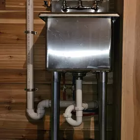 This image displays a freestanding stainless steel utility sink installed against a horizontal wood-paneled wall, likely a Sinks & Sewers LLC project. Exposed PVC drain pipes and supply lines are neatly connected.