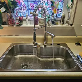 This image captures a stainless steel kitchen sink featuring a newly installed commercial-style pull-down faucet, likely fitted by Sinks & Sewers LLC. Water is running from both the main faucet and sprayer into the basin. The sink sits in a beige countertop below a window.