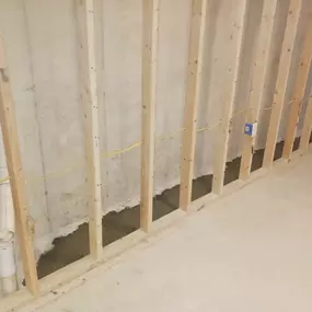 This image displays the interior framing of a basement wall, likely part of a project involving Sinks & Sewers LLC. Wooden studs are visible against the concrete foundation, with rough electrical wiring and a PVC drain pipe stub-out installed.