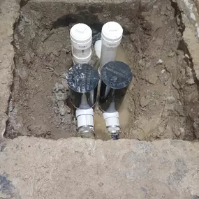 This image shows underground plumbing components exposed in an excavation, likely part of a Sinks & Sewers LLC job. Several pipes, including white PVC and fittings with black 