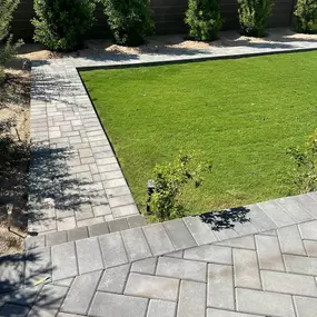 A neatly landscaped backyard with green grass, a block wall, young trees, and modern gray paver walkways in clean patterns.