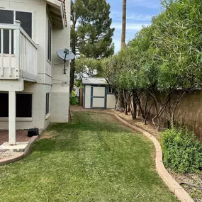 A well-maintained backyard with green grass, trimmed trees, a small shed, and a two-story house featuring a balcony.