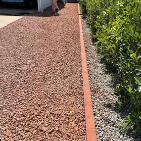 A landscaped side yard with red gravel, a concrete driveway, green shrubs along the edge, and a person walking.