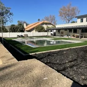 A backyard features a covered pool with artificial grass, modern patio furniture, and neighboring houses under a clear blue sky.