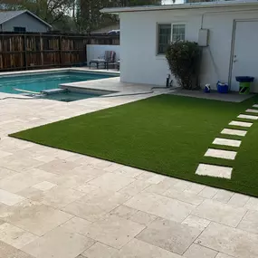 A backyard with a clean patio, a swimming pool, and artificial turf featuring a stepping stone path to a white building.